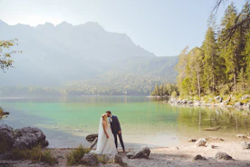 A bride and groom are sharing an intimate moment beside a serene lake, surrounded by lush greenery and distant mountains. The water is clear and reflects the natural beauty of the surroundings.