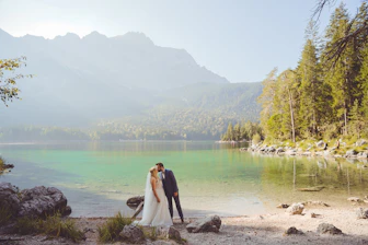 A bride and groom are sharing an intimate moment beside a serene lake, surrounded by lush greenery and distant mountains. The water is clear and reflects the natural beauty of the surroundings.