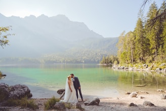 A bride and groom are sharing an intimate moment beside a serene lake, surrounded by lush greenery and distant mountains. The water is clear and reflects the natural beauty of the surroundings.