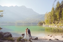A bride and groom are sharing an intimate moment beside a serene lake, surrounded by lush greenery and distant mountains. The water is clear and reflects the natural beauty of the surroundings.