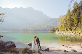 A bride and groom are sharing an intimate moment beside a serene lake, surrounded by lush greenery and distant mountains. The water is clear and reflects the natural beauty of the surroundings.