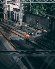 A person wearing an orange safety vest and a hard hat stands on a railway track. The area is surrounded by industrial structures and overhead power lines, with a signal light nearby showing red. The tracks are lined with gravel and the setting is shaded by surrounding walls and vegetation.