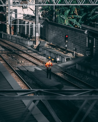 A person wearing an orange safety vest and a hard hat stands on a railway track. The area is surrounded by industrial structures and overhead power lines, with a signal light nearby showing red. The tracks are lined with gravel and the setting is shaded by surrounding walls and vegetation.