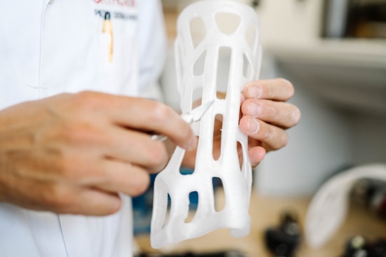 A person in a white lab coat is holding a white, 3D-printed object that resembles a custom orthopedic brace. The object has a lattice structure with several openings. The background suggests a workshop or laboratory environment.