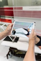 A person is holding a tablet device displaying a digital rendering of a leg with a white medical cast. In the background, there is a medical examination table and glass panels with red stripes.