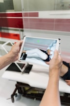 A person is holding a tablet device displaying a digital rendering of a leg with a white medical cast. In the background, there is a medical examination table and glass panels with red stripes.