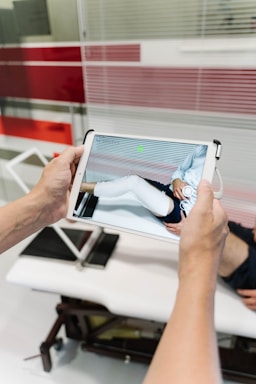 A person is holding a tablet device displaying a digital rendering of a leg with a white medical cast. In the background, there is a medical examination table and glass panels with red stripes.