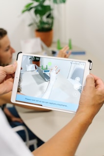 A person holding a tablet, which displays an augmented reality application capturing an image of an arm with digital enhancements overlaid. In the background, a table with a potted plant, documents, and a laptop are visible. Another person is seated and slightly blurred in the background.