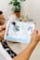 A person holding a tablet, which displays an augmented reality application capturing an image of an arm with digital enhancements overlaid. In the background, a table with a potted plant, documents, and a laptop are visible. Another person is seated and slightly blurred in the background.