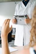 A person wearing a white lab coat is holding a tablet with a specialized handle attachment. Another person's hand is raised in the foreground, suggesting interaction or communication between the two individuals. The scene appears to be in a clinical or professional setting, with blurred backgrounds of furniture and walls.