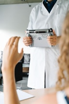 A person wearing a white lab coat is holding a tablet with a specialized handle attachment. Another person's hand is raised in the foreground, suggesting interaction or communication between the two individuals. The scene appears to be in a clinical or professional setting, with blurred backgrounds of furniture and walls.