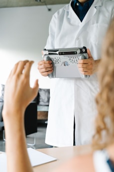 A person wearing a white lab coat is holding a tablet with a specialized handle attachment. Another person's hand is raised in the foreground, suggesting interaction or communication between the two individuals. The scene appears to be in a clinical or professional setting, with blurred backgrounds of furniture and walls.