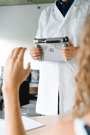 A person wearing a white lab coat is holding a tablet with a specialized handle attachment. Another person's hand is raised in the foreground, suggesting interaction or communication between the two individuals. The scene appears to be in a clinical or professional setting, with blurred backgrounds of furniture and walls.