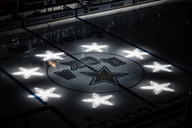 A dimly lit hockey rink featuring a center circle with a star surrounded by illuminated stars. The ice surface reflects soft light, creating a dramatic atmosphere.
