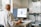 A person wearing a white lab coat is seated at a desk, working on a computer. The computer screen displays what appears to be a digital model or design. The workspace includes a shelf with technical equipment and tools scattered on the desk. A window allows natural light to enter the room, illuminating the scene.