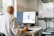 A person wearing a white lab coat is seated at a desk, working on a computer. The computer screen displays what appears to be a digital model or design. The workspace includes a shelf with technical equipment and tools scattered on the desk. A window allows natural light to enter the room, illuminating the scene.