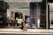 A fashion retail store display with large glass windows showcasing elegant mannequins dressed in upscale clothing. The store interior is brightly lit with a modern, luxurious ambiance. Two people walk past the store, one wearing a long dress and the other a headscarf.