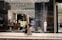 A fashion retail store display with large glass windows showcasing elegant mannequins dressed in upscale clothing. The store interior is brightly lit with a modern, luxurious ambiance. Two people walk past the store, one wearing a long dress and the other a headscarf.