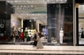A fashion retail store display with large glass windows showcasing elegant mannequins dressed in upscale clothing. The store interior is brightly lit with a modern, luxurious ambiance. Two people walk past the store, one wearing a long dress and the other a headscarf.