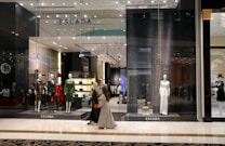 A fashion retail store display with large glass windows showcasing elegant mannequins dressed in upscale clothing. The store interior is brightly lit with a modern, luxurious ambiance. Two people walk past the store, one wearing a long dress and the other a headscarf.