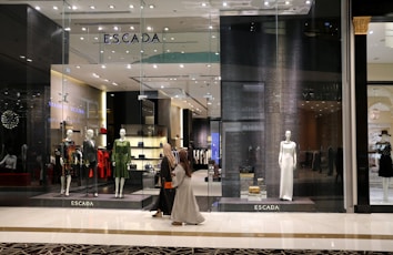 A fashion retail store display with large glass windows showcasing elegant mannequins dressed in upscale clothing. The store interior is brightly lit with a modern, luxurious ambiance. Two people walk past the store, one wearing a long dress and the other a headscarf.