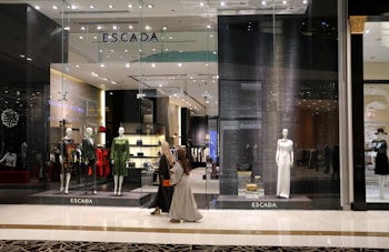 A fashion retail store display with large glass windows showcasing elegant mannequins dressed in upscale clothing. The store interior is brightly lit with a modern, luxurious ambiance. Two people walk past the store, one wearing a long dress and the other a headscarf.