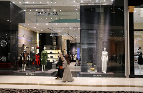 A fashion retail store display with large glass windows showcasing elegant mannequins dressed in upscale clothing. The store interior is brightly lit with a modern, luxurious ambiance. Two people walk past the store, one wearing a long dress and the other a headscarf.