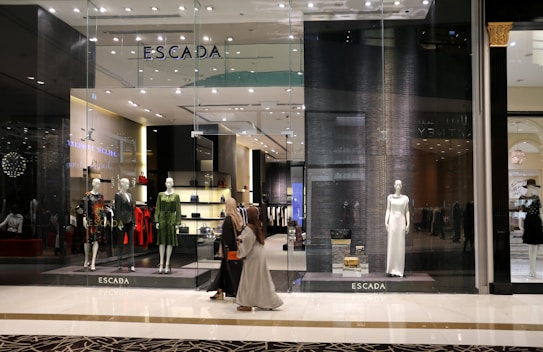 A fashion retail store display with large glass windows showcasing elegant mannequins dressed in upscale clothing. The store interior is brightly lit with a modern, luxurious ambiance. Two people walk past the store, one wearing a long dress and the other a headscarf.