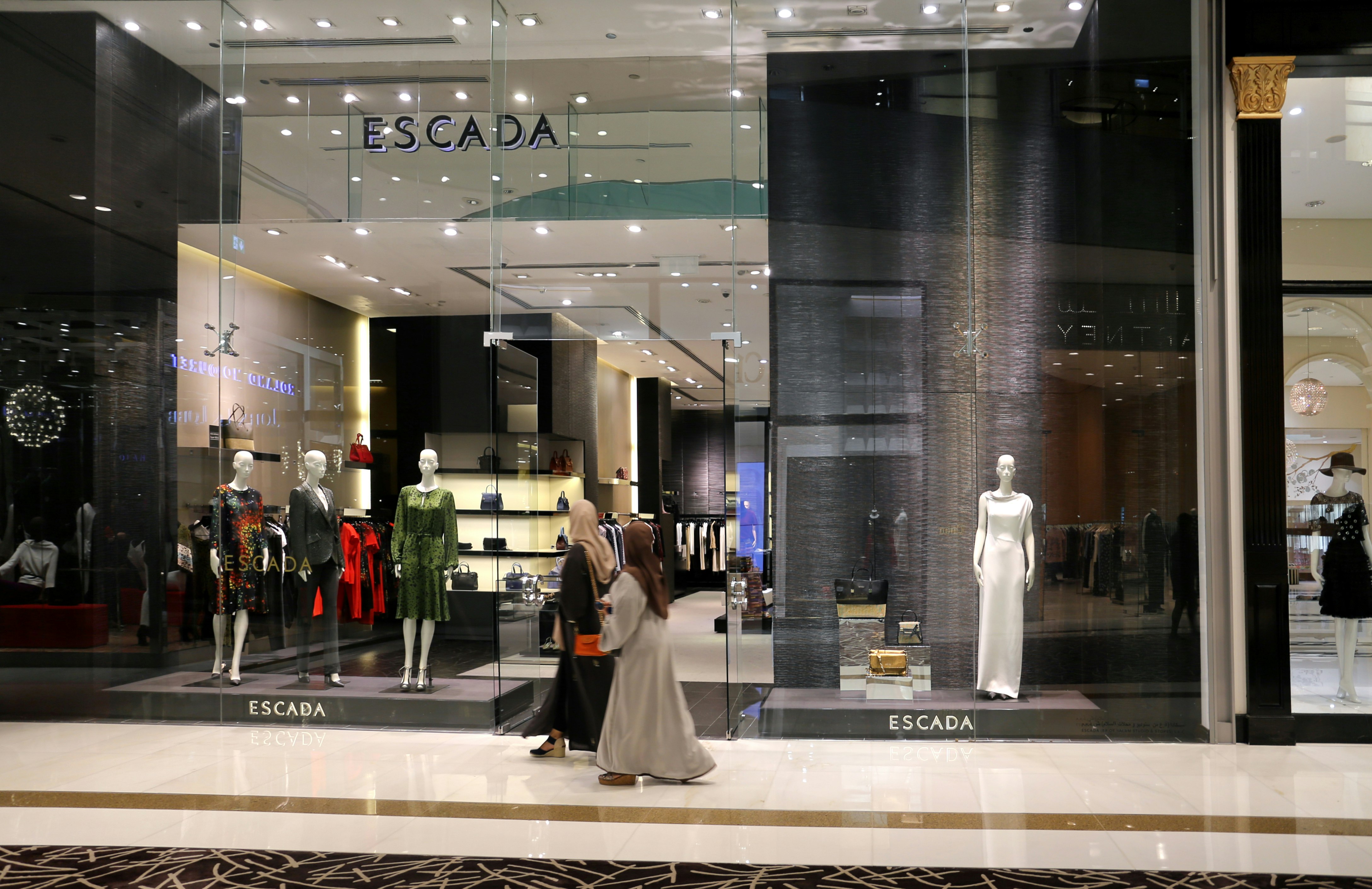 A fashion retail store display with large glass windows showcasing elegant mannequins dressed in upscale clothing. The store interior is brightly lit with a modern, luxurious ambiance. Two people walk past the store, one wearing a long dress and the other a headscarf.