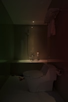 A dimly lit bathroom features modern fixtures including a toilet and a bathtub. A handheld shower head is mounted on the wall. There are towels neatly arranged on a rack and a soap dispenser positioned near the bathtub.