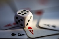A blurred close-up image of a white dice with black dots surrounded by playing cards displaying the ace of hearts and ace of spades.