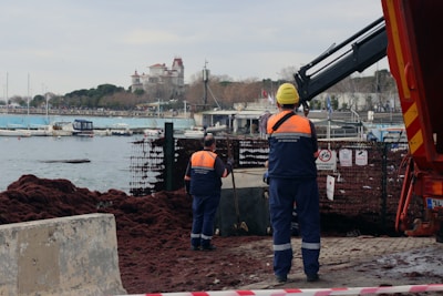 Workers in safety gear, including helmets and high-visibility vests, stand near a waterfront. A crane is present, possibly involved in construction or maintenance work. In the background, there are several boats moored, and a large building with towers is visible across the water. The foreground shows a pile of seaweed or debris.