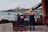 Workers in safety gear, including helmets and high-visibility vests, stand near a waterfront. A crane is present, possibly involved in construction or maintenance work. In the background, there are several boats moored, and a large building with towers is visible across the water. The foreground shows a pile of seaweed or debris.
