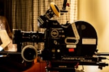 A vintage film camera, specifically an Arriflex 16SR, is positioned on a tripod. The camera features detailed dials, knobs, and labels. The setting includes a blurred curtain backdrop, creating a nostalgic and professional atmosphere.