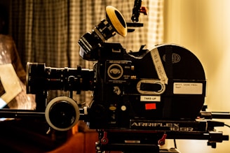 A vintage film camera, specifically an Arriflex 16SR, is positioned on a tripod. The camera features detailed dials, knobs, and labels. The setting includes a blurred curtain backdrop, creating a nostalgic and professional atmosphere.