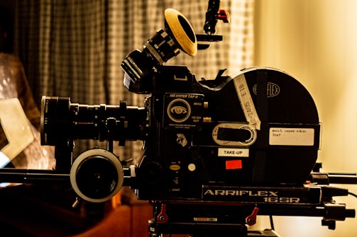 A vintage film camera, specifically an Arriflex 16SR, is positioned on a tripod. The camera features detailed dials, knobs, and labels. The setting includes a blurred curtain backdrop, creating a nostalgic and professional atmosphere.