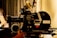 A vintage film camera, specifically an Arriflex 16SR, is positioned on a tripod. The camera features detailed dials, knobs, and labels. The setting includes a blurred curtain backdrop, creating a nostalgic and professional atmosphere.
