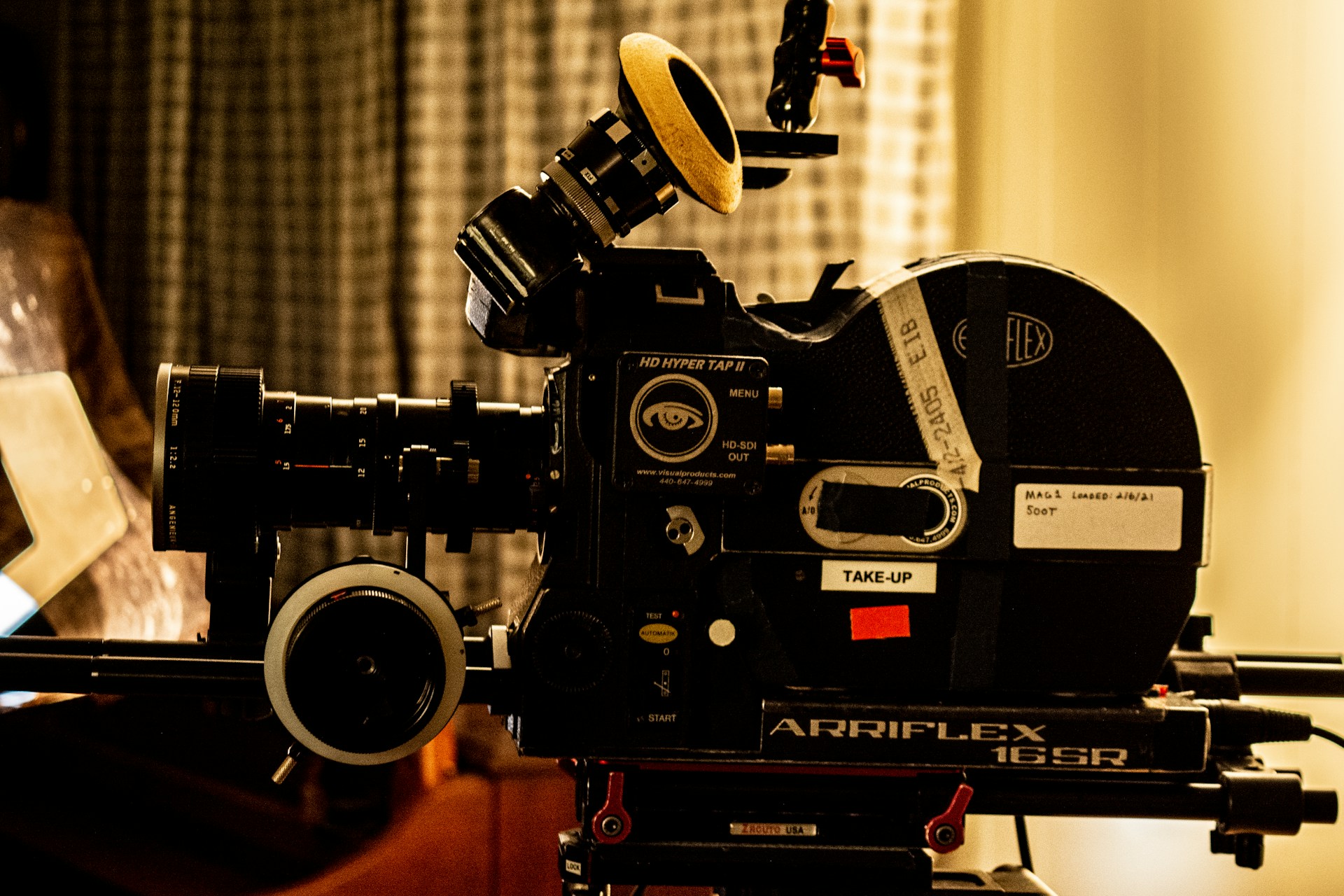 A vintage film camera, specifically an Arriflex 16SR, is positioned on a tripod. The camera features detailed dials, knobs, and labels. The setting includes a blurred curtain backdrop, creating a nostalgic and professional atmosphere.