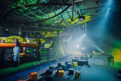 An indoor playground area featuring an inflatable obstacle course with vibrant colors. The play structure resembles a jungle theme with inflatable elements such as trees and an elephant. The surrounding area has foam blocks scattered on the ground and stage lighting casting colorful beams.