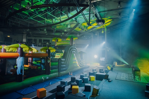 An indoor playground area featuring an inflatable obstacle course with vibrant colors. The play structure resembles a jungle theme with inflatable elements such as trees and an elephant. The surrounding area has foam blocks scattered on the ground and stage lighting casting colorful beams.