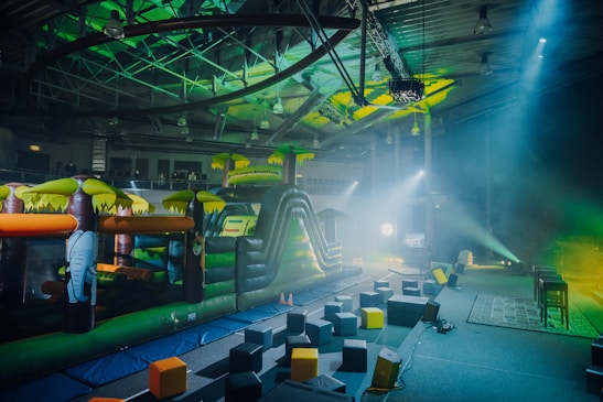 An indoor playground area featuring an inflatable obstacle course with vibrant colors. The play structure resembles a jungle theme with inflatable elements such as trees and an elephant. The surrounding area has foam blocks scattered on the ground and stage lighting casting colorful beams.