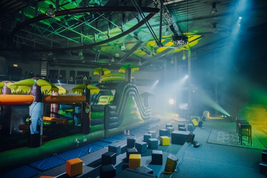 An indoor playground area featuring an inflatable obstacle course with vibrant colors. The play structure resembles a jungle theme with inflatable elements such as trees and an elephant. The surrounding area has foam blocks scattered on the ground and stage lighting casting colorful beams.