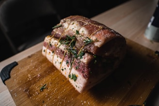 A raw piece of marinated pork is tied with string and garnished with fresh rosemary, resting on a wooden cutting board. The meat is seasoned with herbs and salt, creating a rustic and appetizing appearance.