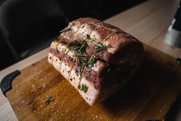 A raw piece of marinated pork is tied with string and garnished with fresh rosemary, resting on a wooden cutting board. The meat is seasoned with herbs and salt, creating a rustic and appetizing appearance.