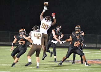 A group of six American football players are actively engaged in a game on a grassy field at night. One player in a white and brown uniform is jumping to catch or block a football. Other players are wearing black and orange uniforms. The scene is lit by bright lights, highlighting the action.