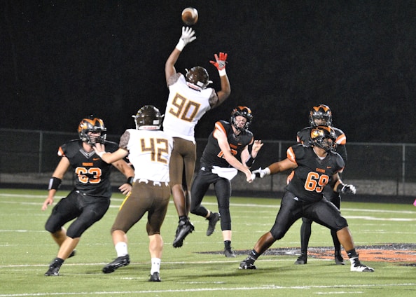 A group of six American football players are actively engaged in a game on a grassy field at night. One player in a white and brown uniform is jumping to catch or block a football. Other players are wearing black and orange uniforms. The scene is lit by bright lights, highlighting the action.