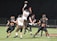 A group of six American football players are actively engaged in a game on a grassy field at night. One player in a white and brown uniform is jumping to catch or block a football. Other players are wearing black and orange uniforms. The scene is lit by bright lights, highlighting the action.