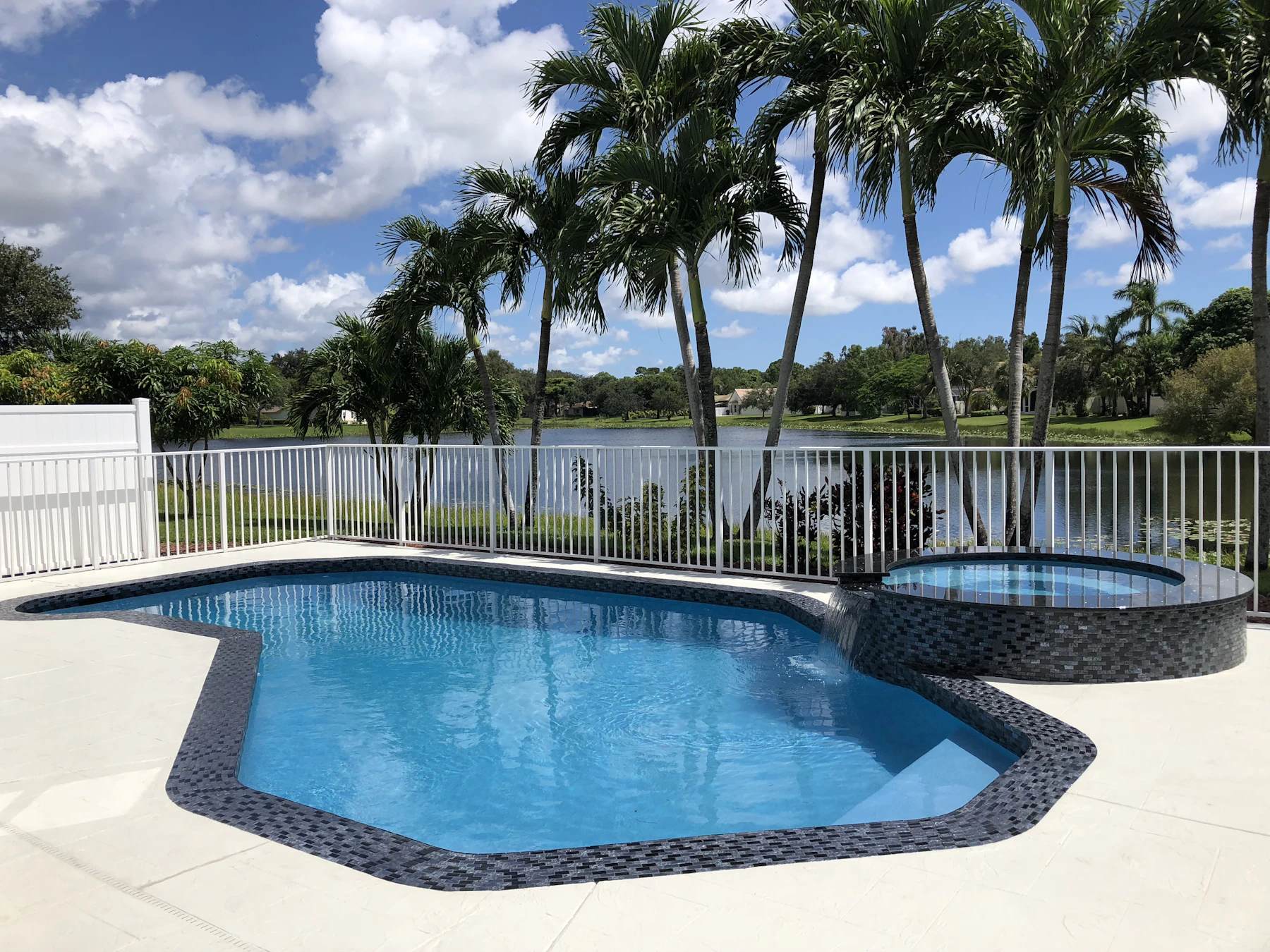 Piscina en forma de riñón y spa elevado con azulejo de vidrio oscuro, palmas y un lago detrás de una cerca de aluminio