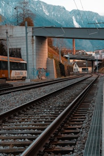 Railway tracks stretch into the distance, bordered by a concrete wall adorned with graffiti. A train is visible on the left side, with the text 'GoldenPass' on it. Above, a bridge crosses over the tracks and snowy mountains create a dramatic backdrop. Vegetation and a chimney are visible on the right, adding to the industrial yet scenic setting.