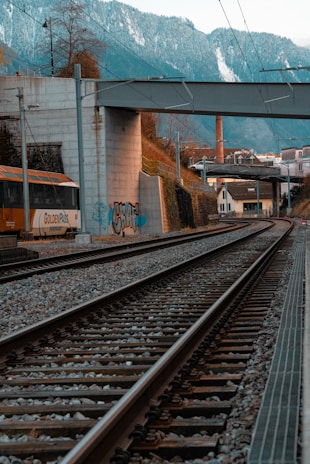 Railway tracks stretch into the distance, bordered by a concrete wall adorned with graffiti. A train is visible on the left side, with the text 'GoldenPass' on it. Above, a bridge crosses over the tracks and snowy mountains create a dramatic backdrop. Vegetation and a chimney are visible on the right, adding to the industrial yet scenic setting.